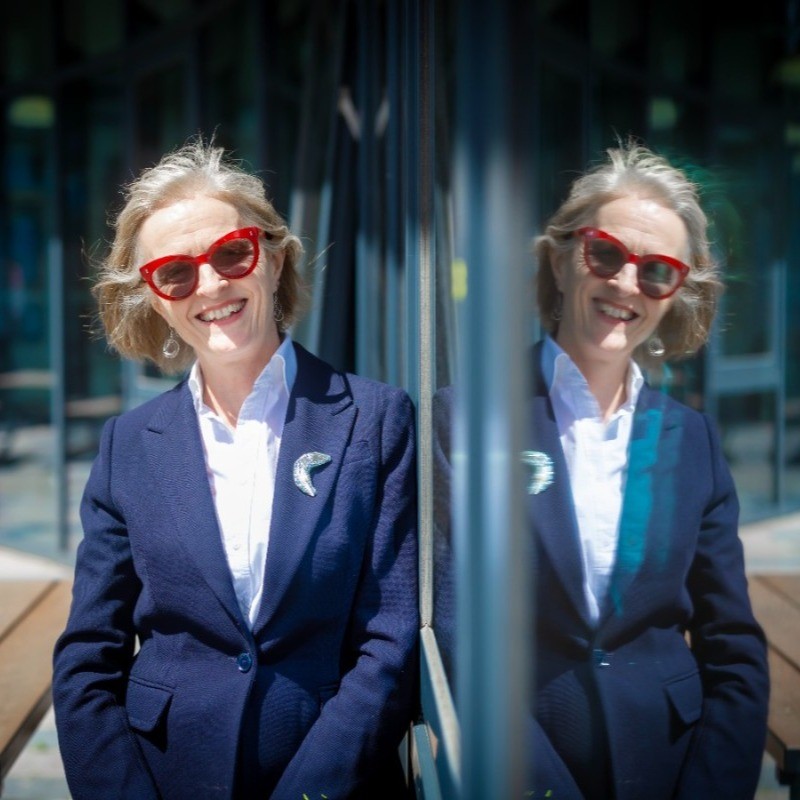 A woman in a blue jacket leans against a glass wall, where her reflection appears