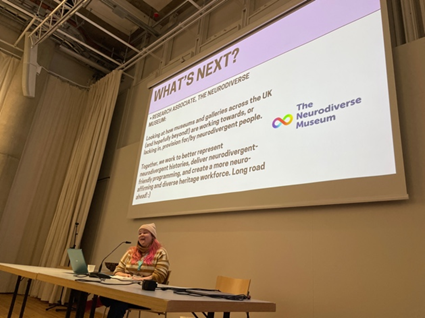 Estonian-UK PhD candidate Iris Sirendi at a long desk speaking to a projection with a slide with the heading &lsquo;What&rsquo;s Next?&rsquo; and a logo that reads &lsquo;The Neurodiverse Museum&rsquo;.