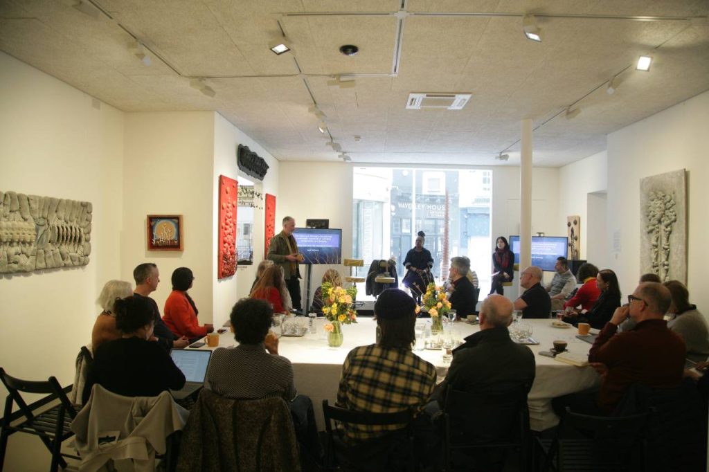 People gathered around a large table looking at someone presenting at a screen. Art is hanging on the walls