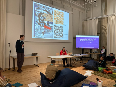  A symposium with person standing to the left holding a microphone; another in the middle, seated, in front of a projection with book cover and QR codes and next to a screen showing live captioning; more people in the foreground on different forms of seating and being;