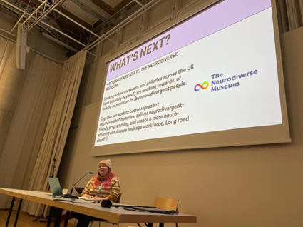 Estonian-UK PhD candidate Iris Sirendi at a long desk speaking to a projection with a slide with the heading ‘What’s Next?’ and a logo that reads ‘The Neurodiverse Museum’.